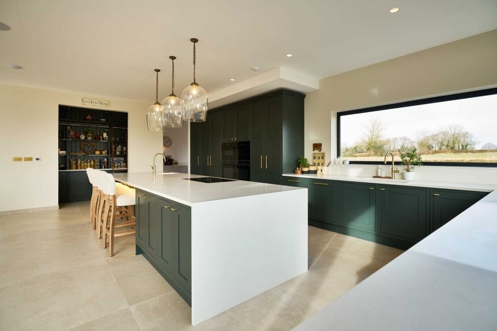 Elegant green shaker kitchen with white waterfall island, brass fixtures, open shelving and countryside views – bespoke design by The Essex Kitchen Design Company 