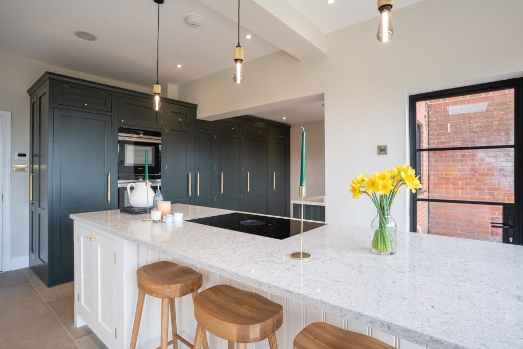 Bespoke green kitchen with dark painted cabinetry, white quartz island, wooden bar stools, and gold accents – designed by The Essex Kitchen Design Company