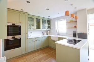 Light green shaker kitchen with white quartz worktops, glass-fronted wall cabinets, double Bosch ovens, a butler sink, and terracotta pendant lighting. 