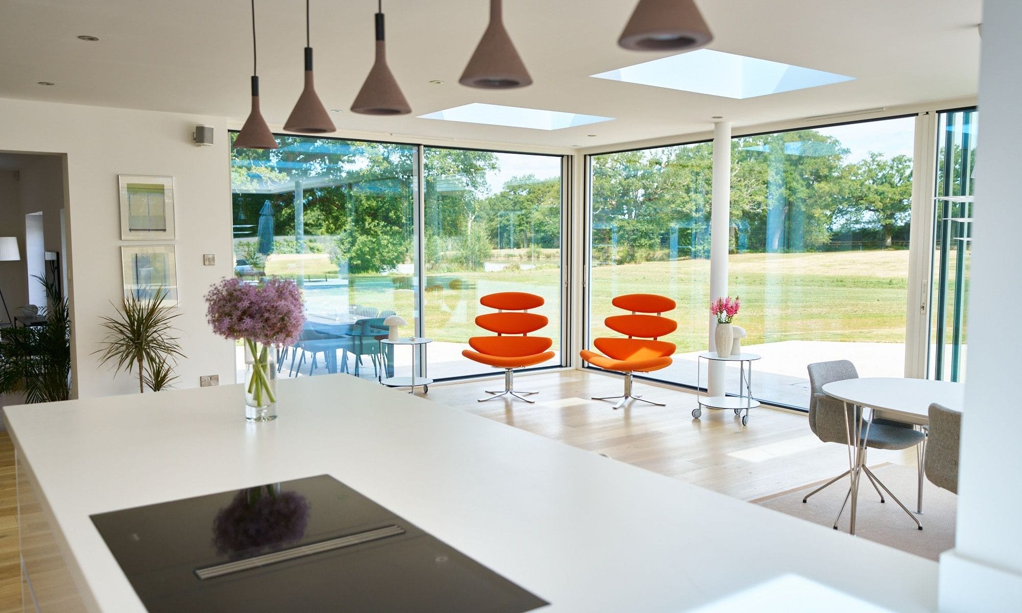 Minimalist handleless kitchen island overlooking full‑height glazing and bright orange lounge chairs.