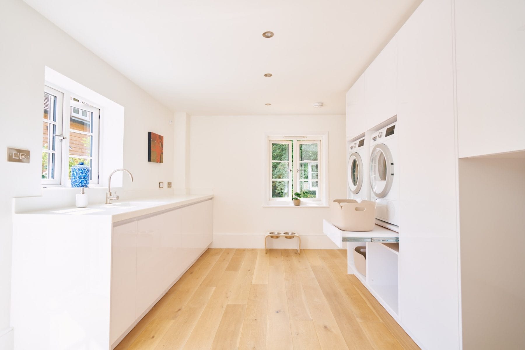 Minimalist handleless kitchen,Light‑filled white gloss utility room with long worktop, stacked laundry appliances and pale oak flooring.
