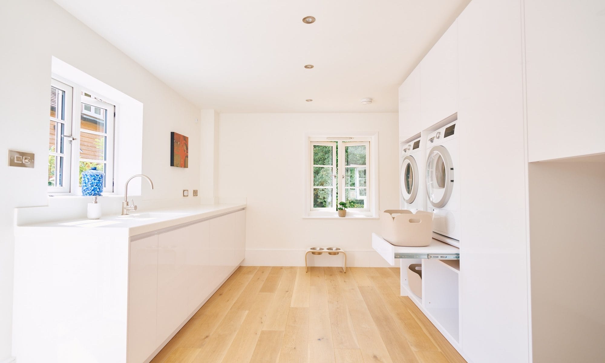 Minimalist handleless kitchen,Light‑filled white gloss utility room with long worktop, stacked laundry appliances and pale oak flooring.